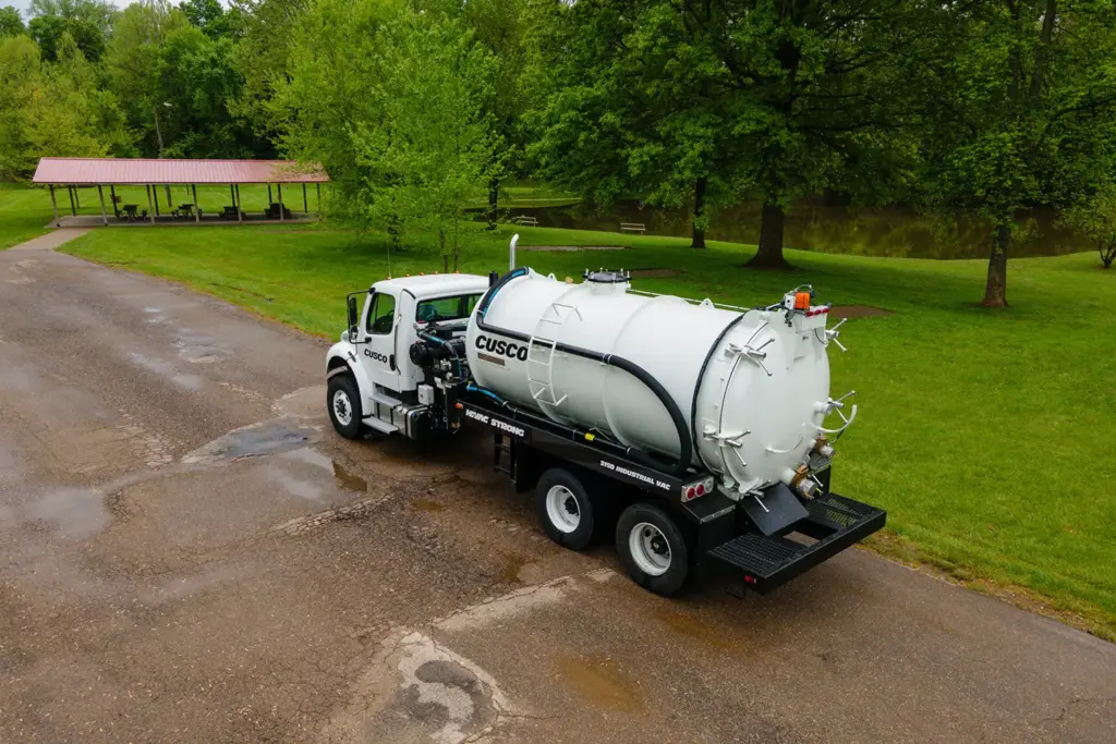 White Cusco vacuum truck with cylindrical tank parked on wet pavement by park pavilion and trees, industrial vacuum equipment